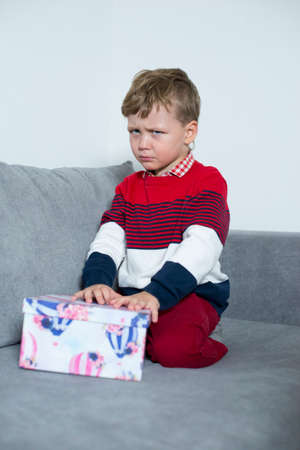 Upset boy sits with a gift on the bed.の写真素材
