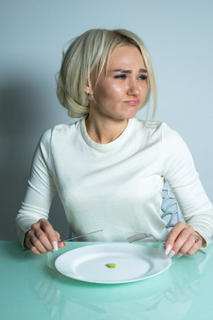 Sick girl with anorexia disease sits at the table and eats diet food. Girl on a diet sits and eats a small portion of food.の写真素材