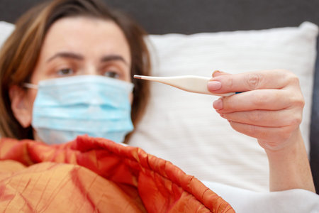 White woman, 30-40 years old, lying on a bed in a medical mask on her face and looking at a...