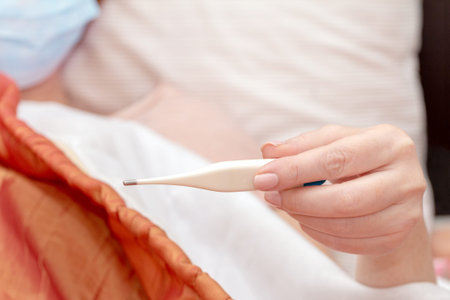 Thermometer in the hand of a woman in a medical mask lying on a bed. sick at home