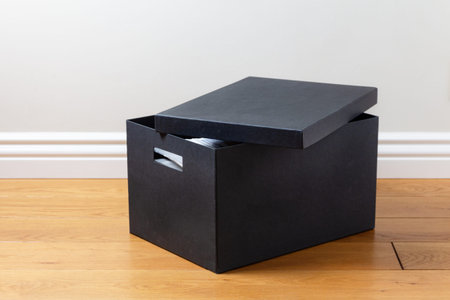 Black cardboard box with ajar lid on a wooden floor near a light wall in the apartment room. Storage system for documents and personal itemsの写真素材