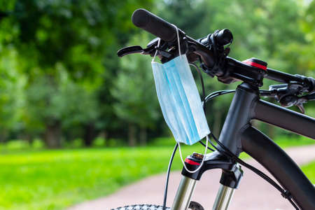 A protective medical mask hangs on a bicycle handlebar against the backdrop of a green park. Coronavirus protection conceptの写真素材