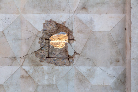 A large round hole with cracks in a reinforced concrete fence with protruding reinforcement rods