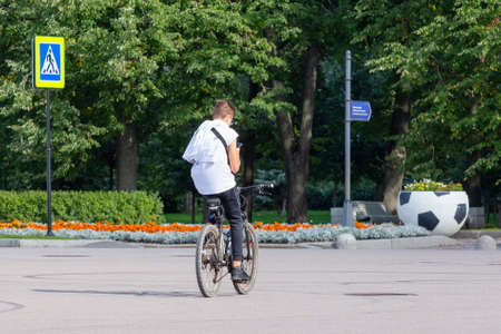 St. Petersburg, Russia - September 04, 2020: Young guy in a white t-shirt and black sports trousers rides a bicycle holding a phone in his hand. Modern urban life - cycling as a sustainable means of transportationのeditorial素材