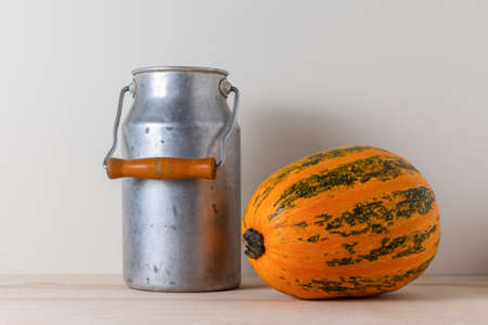 Old aluminum milk can with wooden handle and ripe yellow pumpkin on a kitchen table against a light wall backgroundの写真素材