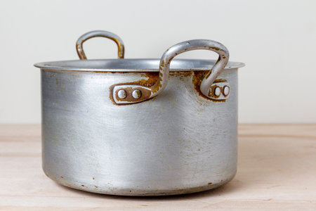 An old rough aluminum saucepan on a wooden kitchen shelf. Used dishes with scratches and marks from...