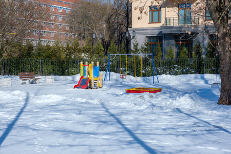 Empty snow-covered playground next to the facade of a modern residential building on a winter sunny dayの写真素材