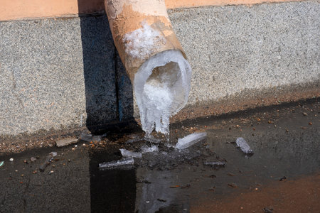 Melting ice and flow of melt water from a drainpipe fixed on the wall of a building outside on a city street in springの写真素材