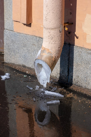 Melting ice and flow of melt water from a drainpipe fixed on the wall of a building outside on a city street in springの写真素材