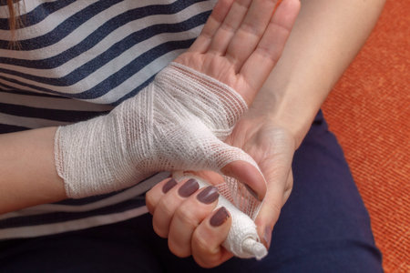 Young woman is bandaging her wounded palm and wrist while sitting on the orange sofa. View from...
