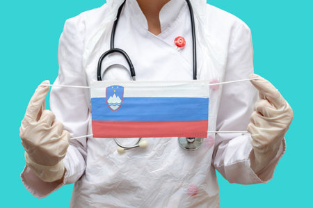 Epidemic in Slovenia. Young woman doctor in a medical coat and gloves holds a medical mask with the print of the flag of Slovenia on a blue background isolated.の写真素材
