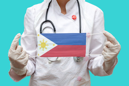 Epidemic in Philippines. Young woman doctor in a medical coat suit and gloves holds a medical mask with the print of the flag of Philippines on a blue background isolatedの写真素材