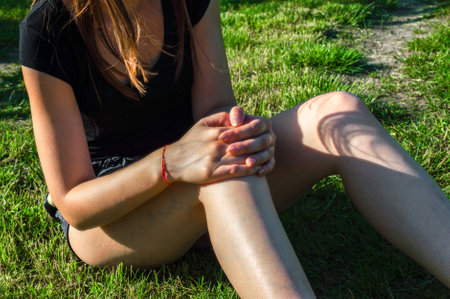 A young woman sitting on a green lawn holds on to a damaged knee. Sprain or cramp Overtrained injured person when training exercising or running outdoorsの写真素材