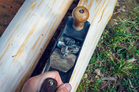 The hand of a young man holds an old manual plane and planes logs outdoors among the green lawn. Top...