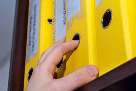 The male hand of an office worker pulls out a folder with company documents from the shelf. On the folders there are inscriptions in Russian: Service contracts, Quality certificates, Holdingの写真素材