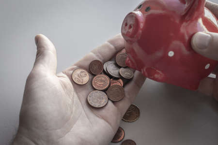 The man holds a red piggy bank and shakes out euro coins into his palm. Gloomy scene. The concept of finding money, crisis, poverty, financial problems, debtsの写真素材