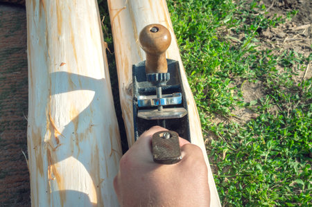 The hand of a young man holds an old manual plane and planes logs outdoors among the green lawn. Top...