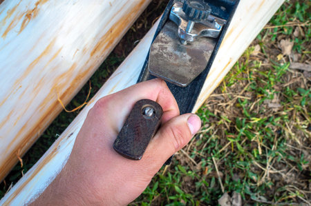 The hand of a young man holds an old manual plane and planes logs outdoors among the green lawn. Top...