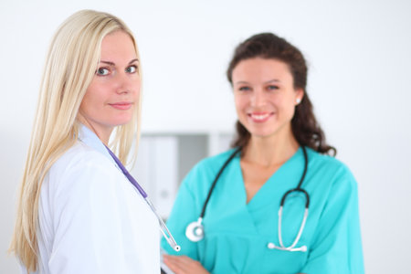 Beautiful young smiling female doctor standing at hospital with doctor on the background. Medical concept.の写真素材