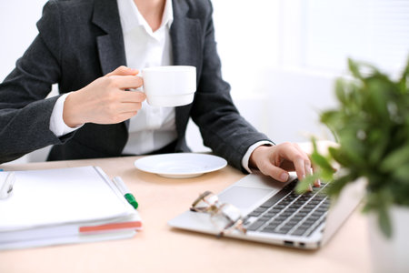Close up of business woman hands with a cup of coffee is sitting at the table and typing on a laptop computer in the white colored officeの写真素材
