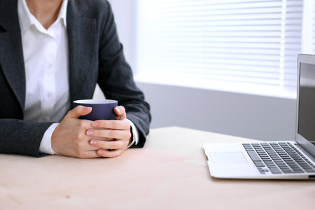 Close up of business woman  hands  with a cup of coffee is sitting at the table and typing on a laptop computer in the white colored office .の写真素材