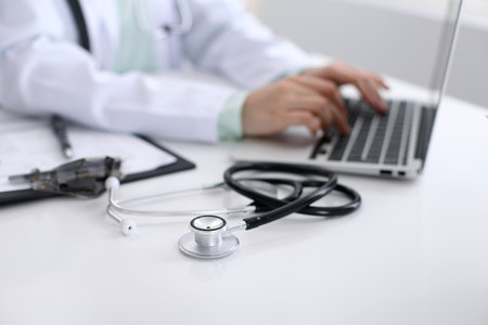 Close-up of stethoscope is lying on the table near female doctor typing on laptop computer.の写真素材