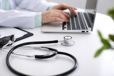 Close-up of stethoscope is lying on the table near female doctor typing on laptop computer.の写真素材