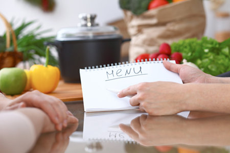 Human hands pointing into note book with copy space area. Two woman making menu in the kitchen, closeup. Cooking and friendship concept.の写真素材