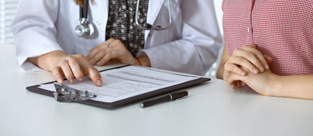 Doctor and patient sitting at the desk. Physician pointing into medical history records and explaining something to young woman. Medicine and health care conceptの写真素材
