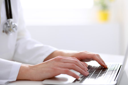 Close-up of female doctor typing laptop sitting at a table in the hospitalの写真素材