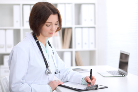Young brunette female doctor is  sitting at the table and  filling up medical application form.の写真素材