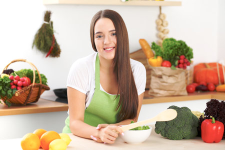 Young brunette woman is cooking or eating fresh salad in the kitchen. Housewife holding wooden spoon in her right hand. Food and health concept.の写真素材