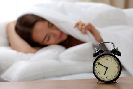 Alarm clock standing on bedside table has already rung early morning to wake up woman in bed sleeping in background. Early awakening, not getting enough sleep, oversleep, time line conceptの写真素材