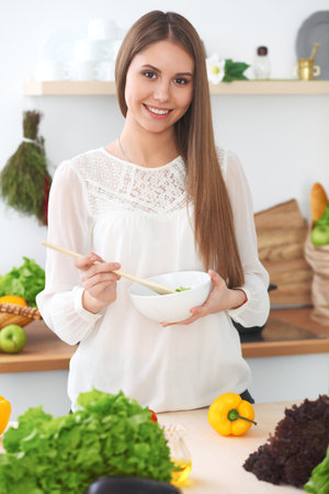 Young happy woman is cooking or eating fresh salad in the kitchen. Food and health conceptの写真素材