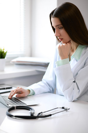 Female doctor brunette sitting  at the table near the window in hospital and typing at laptop computerの写真素材