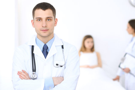 Friendly male doctor on the background with patient in the bed and his physicianの写真素材