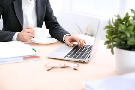Close up of business woman  hands  with a cup of coffee is sitting at the table and typing on a laptop computer in the white colored office .の写真素材