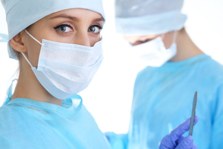 Close up of surgeon woman looking at camera while colleagues performing in background in operation room, isolated. Surgery and medicine conceptの写真素材