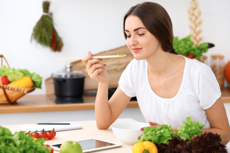 Beautiful  Hispanic woman is cooking in the kitchen. Housewife is tasting fresh salad while sitting at the tableの写真素材