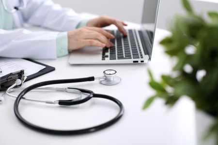 Close-up of stethoscope is lying on the table near female doctor typing on laptop computer.の写真素材