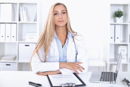 Blonde female doctor sitting at the table in  hospital and looking at cameraの写真素材