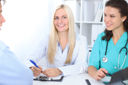 Two confident friendly female doctors sitting at the table and listen to patient . Focus on blonde female doctor. Medical and health care conceptの写真素材
