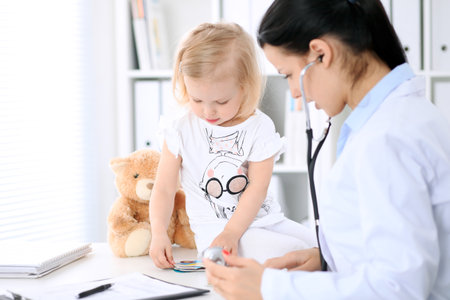 Pediatrician is taking care of baby in hospital. Little girl is being examine by doctor with stethoscope. Health care, insurance and help concept.の写真素材