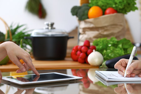 Close-up of  human hands are gesticulate over a tablet in the kitchen. Friends having fun while choosing menu or making online shopping.の写真素材