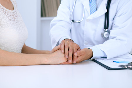 Doctor reassuring his female patient by touching her hands while talking. Symbol of support and trust in medicine.の写真素材