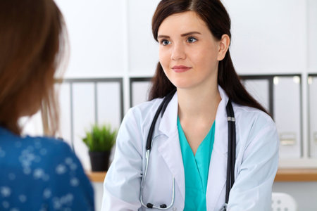 Beautiful female doctor and patient having a talk while sitting at the desk. Physician writes prescription with silver pen at workplace in office. Medicine, pharmacology, prescribing treatment and health care conceptの写真素材