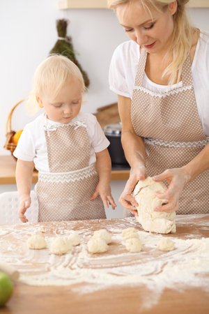 Little girl and her blonde mom in beige aprons  playing and laughing while kneading the dough in the kitchen. Homemade pastry for bread, pizza or bake cookiesの写真素材