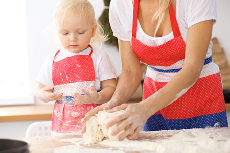 Little girl and her blonde mom in red aprons  playing and laughing while kneading the dough in the...