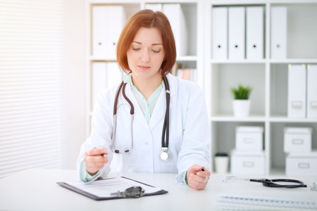 Young brunette female doctor sitting at the table and working at hospital officeの写真素材