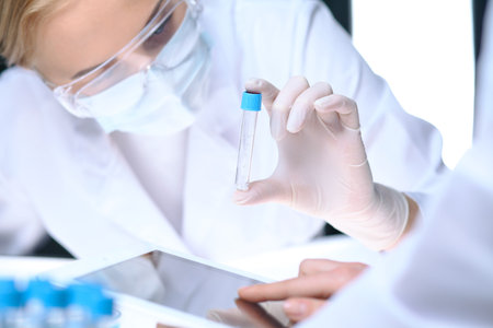Closeup of scientific research team with clear solution in laboratory. Blonde female chemist holds test tube of glass while her colleague checks results with tablet pc. Blood test, medicine or chemical manufacturing conceptの写真素材